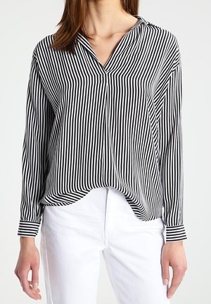 Frau trägt eine langärmlige, schwarz-weiß vertikal gestreifte Bluse, die in hoch taillierte weiße Hosen gesteckt ist, vor einem schlichten Hintergrund.