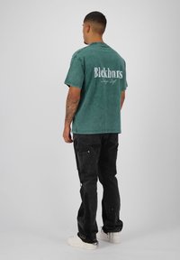 Groen oversized t-shirt met een vervaagde uitstraling, voorzien van witte tekst op de achterkant. Gekocht met zwarte cargobroeken en witte sneakers.