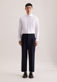 Biała, długa koszula na guziki z delikatnym wzorem, zestawiona z ciemnoniebieskimi spodniami. Model nosi czarne skórzane buty.