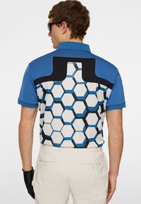 Blå polo skjorta med ett hexagonmönster i svart och ljusblått på vit bakgrund; korta ärmar och klassisk krage.