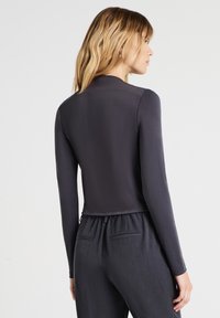 Donna con capelli biondi di lunghezza spalla, che indossa un top aderente a maniche lunghe grigio scuro e pantaloni larghi grigi scuri, che guarda lateralmente voltando le spalle.