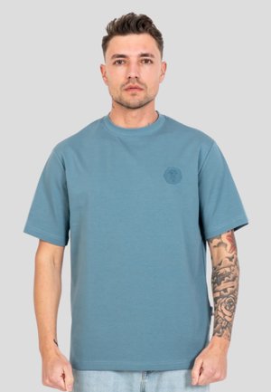 Mand med kort hår iført en ensfarvet blå t-shirt og lyse jeans, der viser en detaljeret tatoveringsærme på sin højre arm mod en ensfarvet baggrund.