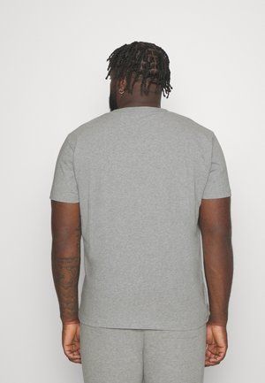 Mann mit kurzen Dreadlocks und Creolen steht mit dem Gesicht zur Wand und trägt ein schlichtes graues T-Shirt und passende graue Hosen.