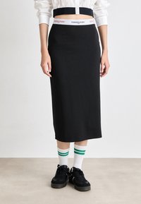 Juodas midi sijonas su Tommy Jeans prekės ženklo baltu juosmeniu, dėvimas su juodais sportbačiais ir baltais kojiniais, kuriuose yra žalių juostelių.