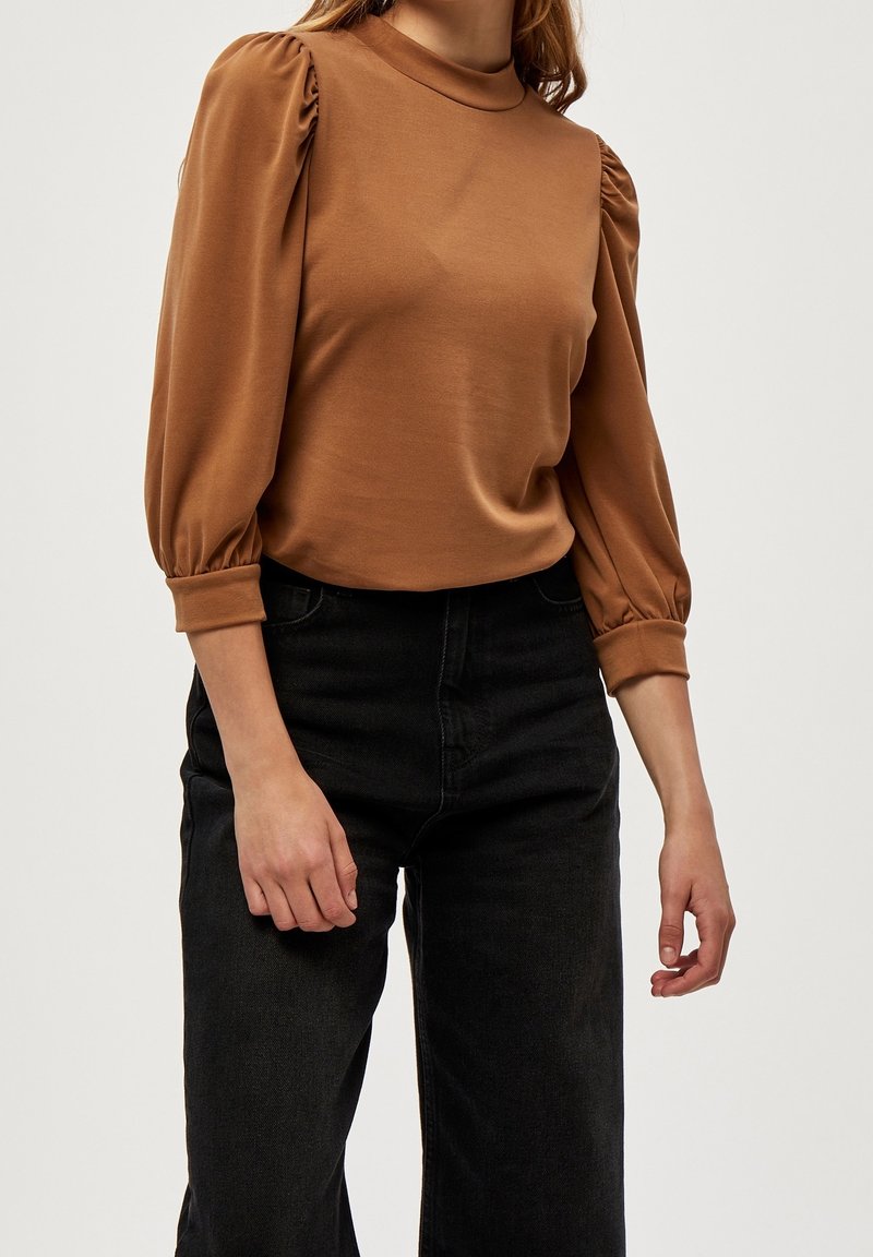 Kvinna i en brun långärmad topp med puffade axlar och svarta högmidjade jeans, stående mot en enfärgad bakgrund.