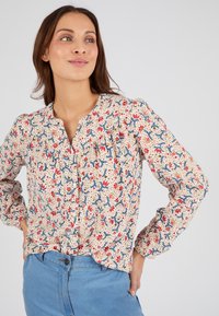 Blusa floreale color crema con motivi botanici rossi e blu. Presenta un colletto rotondo, dettagli arricciati sulle spalle, maniche lunghe e chiusura con bottoni.