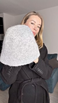 Grå strikket hat med blød tekstur og en afrundet form, holdt af en person i en sort frakke, med et subtilt ribbet mønster.