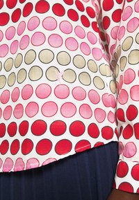 Kortärmad blus med vit bakgrund, prydd med flerfärgade cirklar i röd, rosa och grön. Märkbar sömdetalj och nederkant.