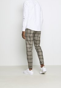 En person som bär beige och grå rutiga byxor, en vit långärmad skjorta och vita Adidas-sneakers står mot en enkel vägg.