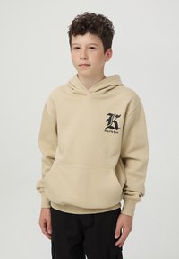 Felpa beige con tasca a marsupio, cappuccio regolabile con cordino e logo nero sul lato sinistro del petto. Tessuto con texture e polsini a coste.