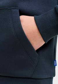 Sudadera azul marino oscuro con bolsillo frontal. La tela es suave con puños acanalados, destacando un diseño limpio y moderno. Etiqueta azul visible.