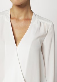 Weiße Bluse mit V-Ausschnitt, langen Ärmeln und einem leichten, glatten Stoff. Auffälliges goldenes Reißverschlussdetail am Ausschnitt.