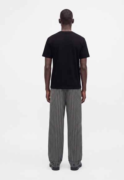 Homme debout de dos, portant une chemise noire à manches courtes et un pantalon large gris à rayures verticales avec des chaussures noires.