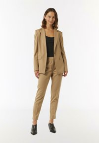 Blazer beige e pantaloni aderenti coordinati in tessuto liscio, abbinati a un top nero. Modella con scarpe col tacco nere posata su uno sfondo bianco.