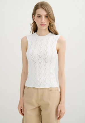 Mujer con cabello rubio largo que lleva una camiseta de punto blanca sin mangas con un patrón de hojas y pantalones beige, de pie frente a un fondo liso.