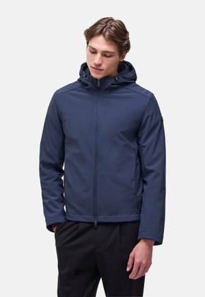Giovane uomo che indossa una giacca blu navy con cappuccio e zip, con le mani nelle tasche, che guarda verso il basso, su uno sfondo chiaro e uniforme.