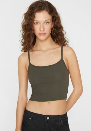 Junge Frau mit lockigem braunem Haar, die ein olivgrünes bauchfreies Oberteil und schwarze Jeans trägt und vor einem weißen Hintergrund steht.