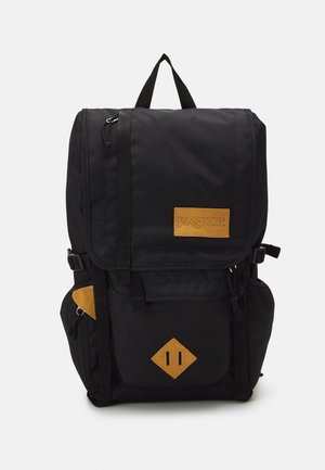 Sac à dos en tissu noir avec des accents en cuir. Il dispose d'un rabat supérieur avec des sangles ajustables, de poches latérales et d'une patch en forme de diamant à l'avant.