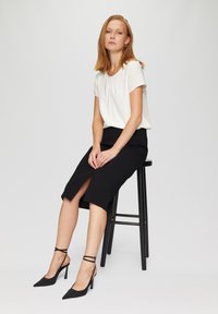 Weiße kurzärmlige Bluse kombiniert mit einem schwarzen kniebombierenden Bleistiftrock mit Schlitz vorne und schwarzen Stilettos mit Knöchelriemen.