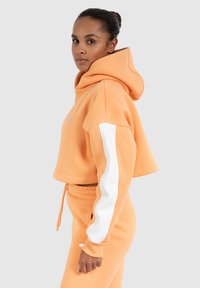Korte oranje hoodie met witte zijstrepen, met een trekkoord, zachte textuur en een klein logo aan de voorkant. Draagt met bijpassende broek.