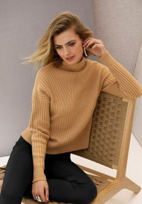 Eine Frau mit blonden Haaren, die einen beigen gerippten Pullover und schwarze Hosen trägt, sitzt auf einem geflochtenen Holzstuhl und passt einen langen Perlenohrring an.
