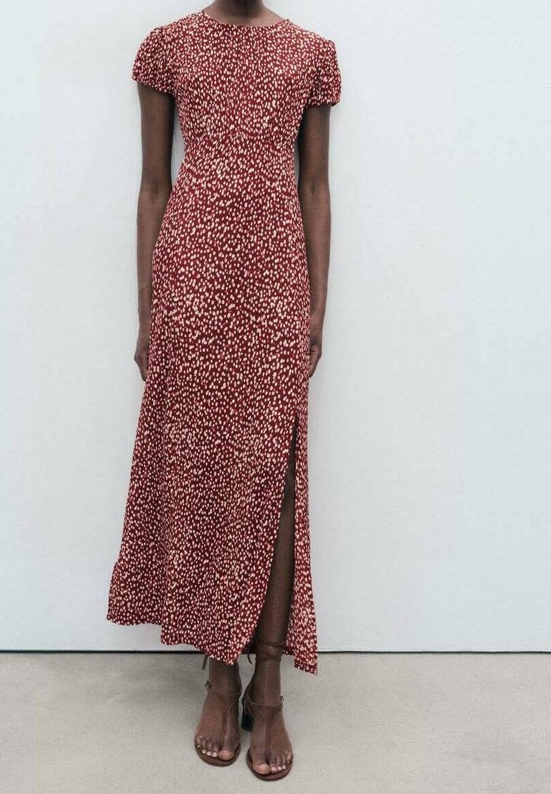 Femme portant une robe midi rouge à pois blancs, manches courtes, fente latérale, et des sandales marron à talons blocs avec bride à la cheville, debout contre un mur blanc.
