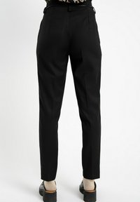 Osoba w dopasowanych czarnych spodniach, beżowych skarpetkach i czarnych butach na platformie, stoi na tle jednolitego białego tła, widok z tyłu.