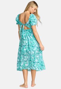 Une robe midi florale teal avec des manches bouffantes, un dos ouvert maintenu par un lien, et une jupe fluide avec une coupe ample et une texture douce.