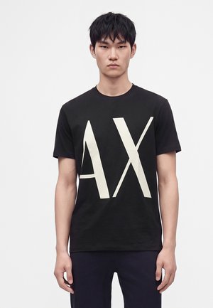 Jeune homme aux cheveux courts et noirs, portant un t-shirt noir avec de grandes lettres blanches "AX" à l'avant sur un fond uni.