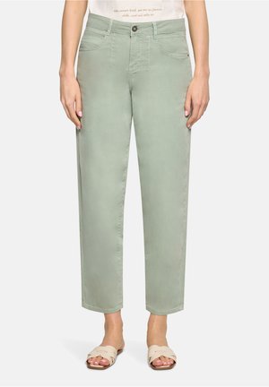 Kvinde iført lysegrønne, lige, cropped bukser med forlommer og beige slidesandaler, stående mod en ensfarvet baggrund.