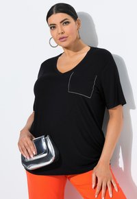 Schwarzes Kurzarmoberteil mit V-Ausschnitt, ausgestattet mit einer dekorativen Tasche, die mit Strasssteinen umrandet ist. Kombiniert mit orangen Hosen und einer metallisch silbernen Tasche.