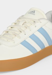 Λευκό αθλητικό παπούτσι Adidas με ανοιχτομπλε ρίγες, μπεζ κορδόνια, καουτσούκ σόλα και λογότυπο Adidas στη γλώσσα.