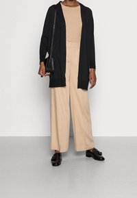 Cárdigan largo negro sobre un top beige acanalado y pantalones beige de pierna ancha, combinados con mocasines negros y un pequeño bolso cruzado negro.