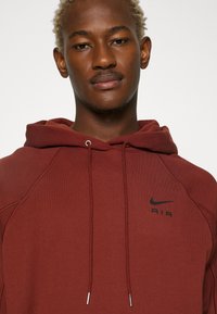 Bordeaux Nike-hoodie med snörning, med en svart "AIR"-logotyp på bröstet och raglanärmar. Slät tygtextur, avslappnad design.