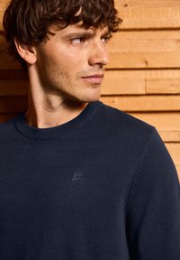 Marineblau gestrickter Pullover mit Rundhalsausschnitt, strukturiertem Stoff und einem kleinen Logopatch auf der Brust. Hintergrund aus Holzpaneelen.