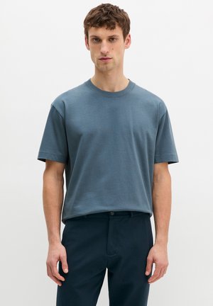 Junger Mann mit kurzem braunem Haar, trägt ein gedämpft blaues, kurzärmeliges Rundhals-T-Shirt und dunkelblaue Hose, steht vor weißem Hintergrund.