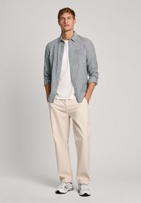 Camisa de cuadros azul y blanca sobre una camiseta blanca, combinada con pantalones beige claros y zapatillas gris-blancas. Fondo liso.