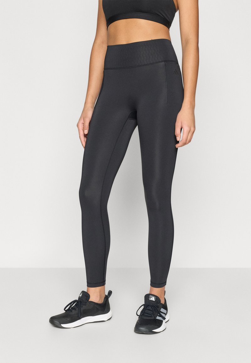 Leggings de sport noirs avec une taille haute, une texture lisse et un détail de poche latérale, assortis à des chaussures de sport noires.