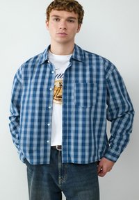 Camicia a quadri blu e bianchi con colletto button-down, maniche lunghe e una tasca sul petto, indossata sopra una t-shirt bianca con grafica.