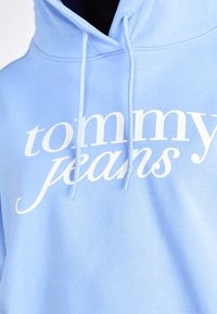 Sudadera de color azul claro hecha de tela suave, con el texto del logotipo en blanco "tommy jeans" en el pecho, y cordones en la capucha.
