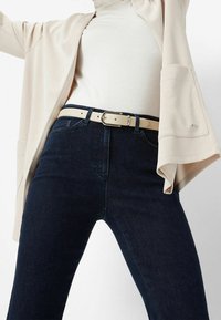 Cardigan abierto beige sobre una blusa blanca, combinado con jeans de cintura alta azul oscuro y un cinturón beige claro con hebilla de plata.