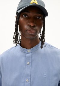 Hellblaues Button-Down-Hemd mit Mandarinkragen, versehen mit einem kleinen gestickten Logo und zwei Knöpfen am Kragen. Getragen mit einer grauen Mütze.