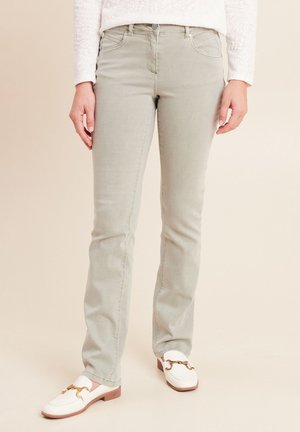 Frau trägt hellgraue Slim-Fit-Jeans und weiße Mokassins mit goldenen Schnallen, stehend vor einem schlichten neutralen Hintergrund.