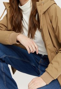 Personne portant une veste à capuche beige, un col roulé blanc et un jean bleu, assise les jambes croisées et les mains reposant sur le genou et la cuisse.