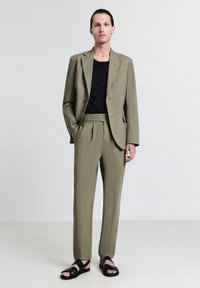 Completo verde oliva con giacca su misura e pantaloni plissettati, abbinato a una maglietta nera e completato da sandali neri a scivolo.