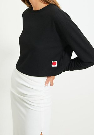 Vrouw draagt een zwart cropped top met lange mouwen en een klein rood appelschildje en een witte geplooide rok, hand rustend op haar heup.