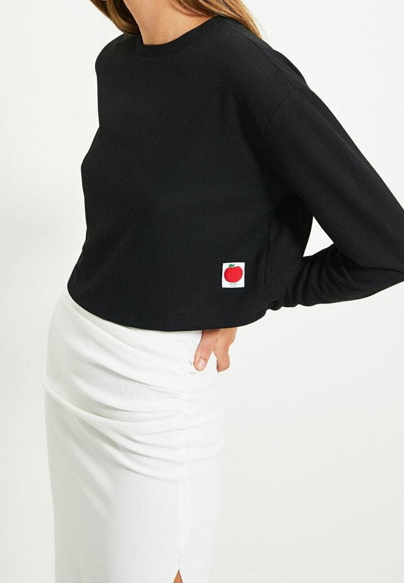 Femme portant un crop top noir à manches longues avec une petite écusson pomme rouge et une jupe froncée blanche, main reposant sur la hanche.