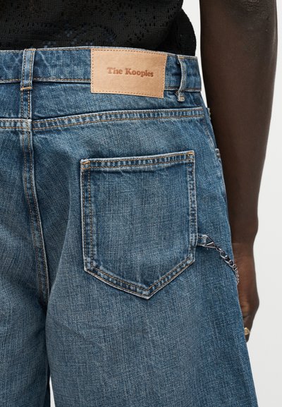 Πίσω όψη τζιν παντελονιών από denim με δερμάτινη ετικέτα μάρκας "The Kooples", ορατή πίσω τσέπη και αλυσίδα σαν αξεσουάρ που κρέμεται από το πλάι.