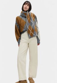 Pull en tricot à losanges marron, gris et blanc avec un col haut, associé à un pantalon large crème et des chaussures beige. Tissu texturé avec une apparence chaleureuse.
