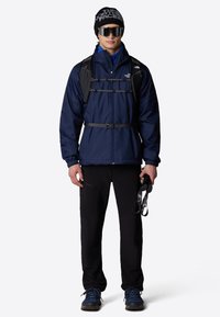 Merijalkatakki säädettävillä hihnoilla, mustat housut, siniset vaelluskengät ja musta pipo. Hyväilee vaellussauvoja, pä�ässään suuret aurinkolasit.